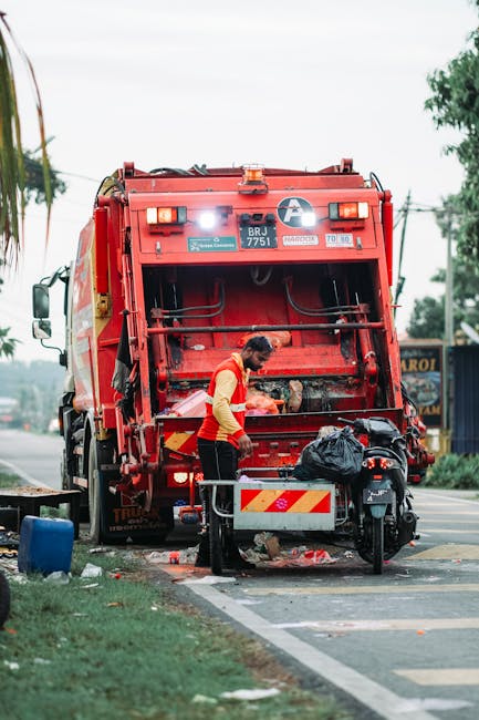 A street scene featuring a worker operating a red waste collection vehicle, which is positioned on the roadside with its rear hatch open, revealing the loading mechanism. The worker, dressed in a yellow and red safety vest, is standing behind a small metal cart or bin, emptying items into the vehicle. The vehicle's exterior is red with reflective stripes and identification labels, including a white circular logo and a vehicle registration number. Nearby, a black motorcycle is parked beside the worker, with a black garbage bag securely placed on its seat. In the background, a sidewalk with some greenery and trees, along with a few blurred storefronts or signs, can be seen. The scene is illuminated by natural daylight under a slightly overcast sky, typical of a thoroughfare where private waste collection or rubbish removal services are regularly conducted, reflecting an alternative approach to municipal collection. Waste Disposal Edgware occasionally operates in such settings, providing on-site clearance and waste handling outside standard local authority services.