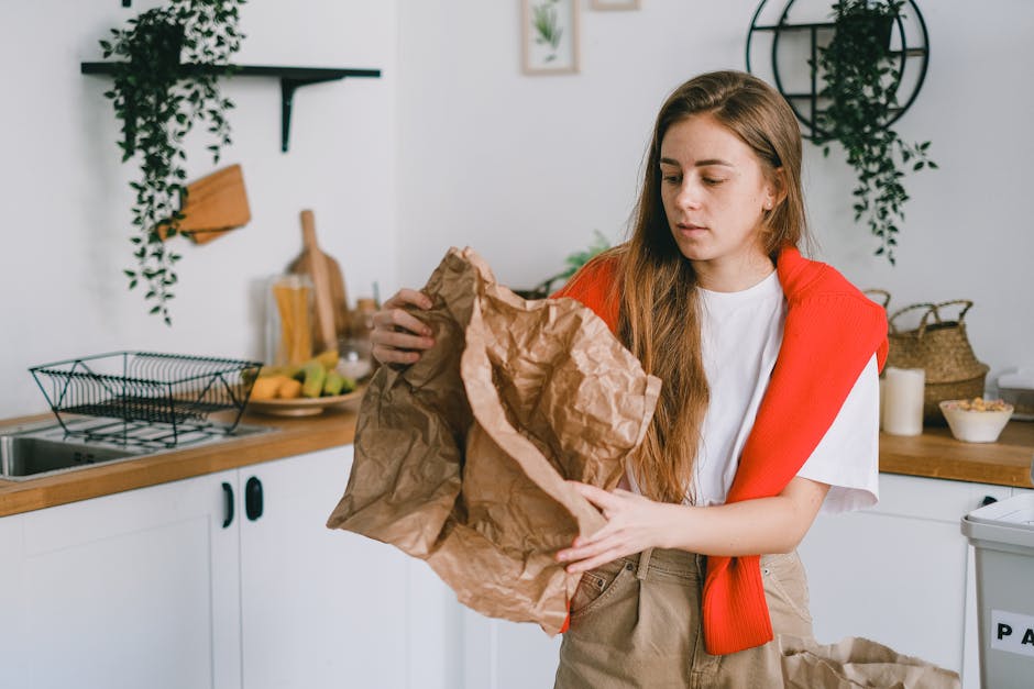 A young woman with long, light brown hair is standing in a modern kitchen, holding a large crumpled brown paper bag with both hands and examining its contents. She is wearing a white T-shirt with a red sleeveless sweater draped over her shoulders, and beige pants. The background features white cabinets with black handles, a wooden countertop, and various kitchen items including a wire dish rack, a bowl of fruit, and a cutting board, with a few potted plants and framed artwork on the wall. The lighting is natural and bright, and the overall scene suggests she is preparing to dispose of waste or recyclable materials, aligning with the theme of rubbish removal or household waste management. The setting is clean, organized, and indicative of a domestic environment where waste sorting or disposal may be occurring, reflective of private waste handling services such as those provided by Waste Disposal Edgware.