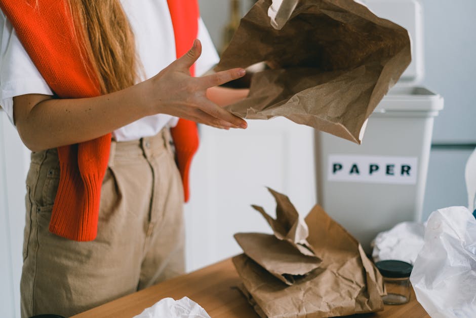 A young woman with long brown hair, wearing a white t-shirt and a red vest, is standing at a kitchen countertop, holding a crumpled brown paper bag and examining it. The countertop is cluttered with various crumpled paper sheets, plastic wrappers, and containers, indicating waste materials. To her right, there is a white recycling bin labeled 'PAPER' with its lid open, and nearby are transparent plastic bottles and a blue and pink plastic container for eggs or similar items. In the background, the kitchen features a white backsplash, a wooden countertop with a stainless steel sink and faucet, and decorative green plants hanging on the white wall. The scene is well-lit with natural or artificial light, reflecting a typical household setting involved in waste disposal or rubbish sorting activities, possibly aligned with private rubbish collection or disposal services offered by Waste Disposal Edgware.