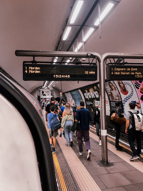 A busy underground railway platform at Edgware Tube Station, showing a group of passengers waiting and walking along the platform. The scene includes various individuals dressed in casual and business attire, some carrying backpacks or shoulder bags. The platform has a textured yellow safety line near the edge, with advertising posters and station signage visible along the walls and columns. Overhead, illuminated digital displays provide train information, including destinations such as Morden and High Barnet, with approximate arrival times. The station's ceiling is modern, featuring exposed structural elements and bright lighting fixtures. The environment appears clean and well-maintained, reflecting the typical setting for a London-area underground station, supporting the context of transport and travel related to local public transportation services. Waste Disposal Edgware is not directly related to this scene but might handle excess waste generated by station upkeep or commuters' discarded items during their journey.