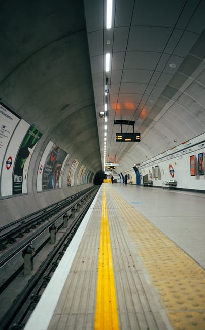 A view of an underground train station platform with a curved ceiling covered in grey panels, illuminated by a series of long, narrow ceiling lights. On the left side, there are train tracks with a gap between the platform edge and the rails, marked by a yellow tactile paving strip for safety. To the right, there are advertising posters attached to the white tiled wall, some displaying brands and promotional content. The platform floor includes a textured yellow strip and a smooth grey surface. At the far end of the station, a digital information display board shows train timings in bright yellow text, with several blurred figures of commuters standing or walking in the background. The overall scene appears clean and well-maintained, characteristic of a modern urban transit environment, where public transportation infrastructure supports passenger movement in the city. Waste Disposal Edgware occasionally handles waste removal at such facilities, ensuring proper disposal of waste generated in and around transit stations, although no specific waste-related items are visible in this image.