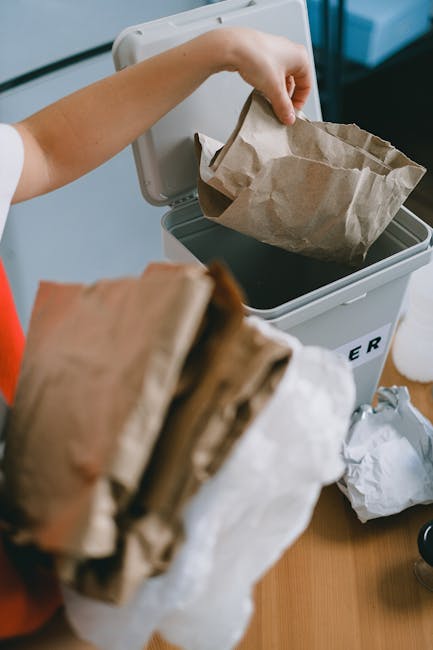 A person is shown disposing of a crumpled brown paper bag into a white indoor rubbish bin, which has a lid partially open. The bag appears used and is being lifted from a pile of similar paper waste held in the person's hand, with some paper waste resting on a nearby surface. The waste bin, made of plastic, is situated on a wooden surface and is designated for general rubbish collection. The background is blurred, but hints of an indoor environment are visible. This scene illustrates a typical household or office setting where independent waste disposal and rubbish removal services by Waste Disposal Edgware may be employed for on-site waste management or alternative collection options, emphasizing environmental responsibility and clean disposal practices.