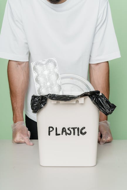 A person wearing a white t-shirt and transparent disposable gloves is standing behind a white plastic rubbish bin labeled 'PLASTIC' in black letters. The bin is filled with various plastic waste materials, including a clear plastic egg carton and a crumpled black plastic bag draped over the edge. The background is a plain light green wall, and the scene is lit evenly, emphasizing the focus on plastic waste disposal. This setup exemplifies responsible plastic waste separation, aligning with private rubbish collection or alternative waste handling services provided by Waste Disposal Edgware for efficient and environmentally conscious rubbish removal in the HA8 Edgware area.