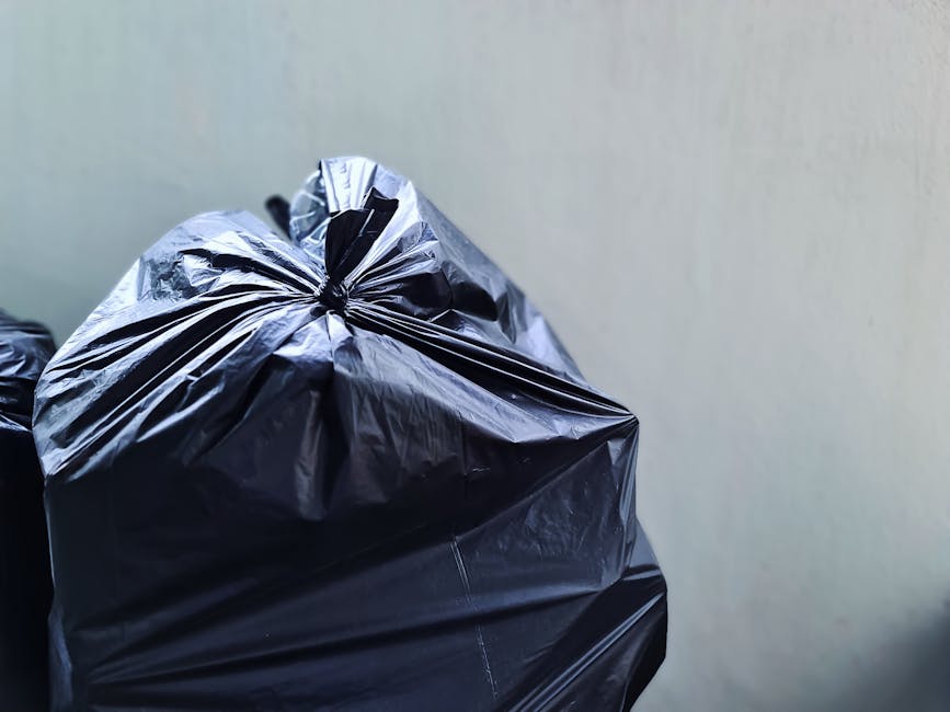 A person is shown disposing of a crumpled brown paper bag into a white indoor rubbish bin, which has a lid partially open. The bag appears used and is being lifted from a pile of similar paper waste held in the person's hand, with some paper waste resting on a nearby surface. The waste bin, made of plastic, is situated on a wooden surface and is designated for general rubbish collection. The background is blurred, but hints of an indoor environment are visible. This scene illustrates a typical household or office setting where independent waste disposal and rubbish removal services by Waste Disposal Edgware may be employed for on-site waste management or alternative collection options, emphasizing environmental responsibility and clean disposal practices.
