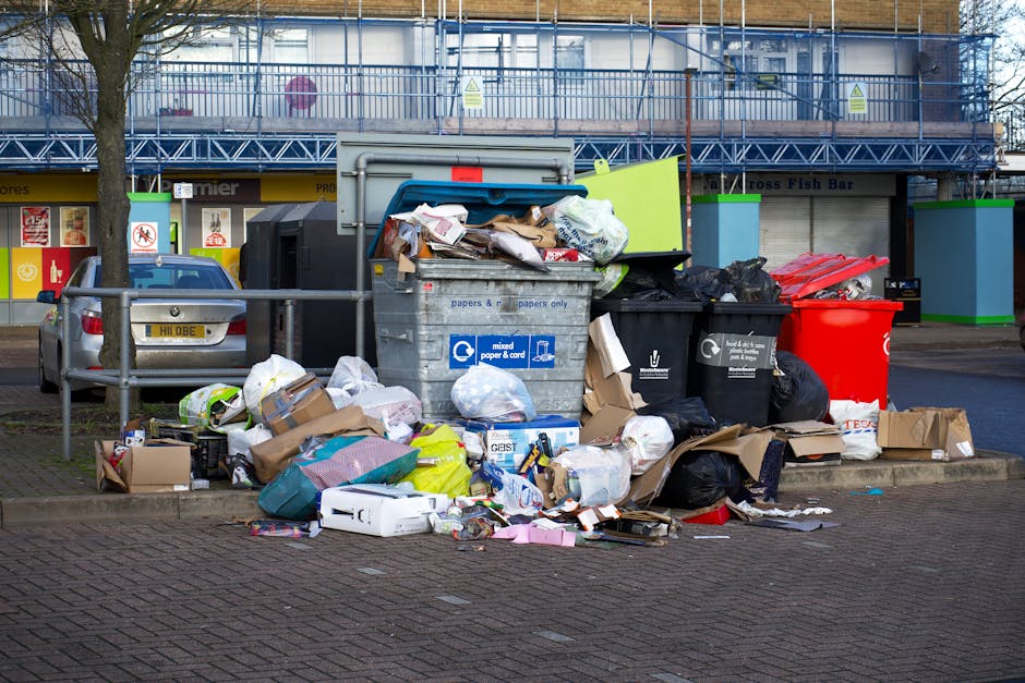 In a designated parking area, a large collection of overflowing waste bins and accumulated rubbish is visible, including a grey mixed paper and card bin with its lid open and contents spilling out. Surrounding this bin are various plastic bags, some black and some white, filled with waste, along with flattened cardboard boxes, paper packaging, and assorted litter scattered on the brick-paved ground. To the left, there is a metal barrier fence and a parked grey car with a UK registration plate. In the background, a storefront with closed shutters and signage is visible, along with a building under construction or renovation, wrapped in scaffolding and blue protective netting. The scene appears to be outdoors on a street, possibly representing an example of improperly managed rubbish collection or excess waste typical of private disposal services like Waste Disposal Edgware, rather than regular council curbside collection. The environment is illuminated by natural daylight, emphasizing the chaotic accumulation of household or commercial waste.