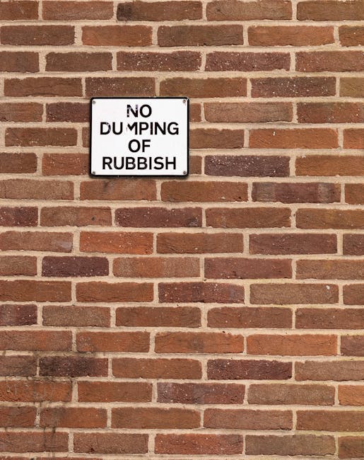 A rectangular white metal sign with bold black text reading 'NO DUMPING OF RUBBISH' is affixed to a red brick wall. The wall features a traditional running bond pattern with reddish-brown and darker mortar joints, providing a textured background. The sign is centered in the upper portion of the image, slightly above middle height, and appears to be attached with four screws, one at each corner. Natural daylight illuminates the scene, casting soft shadows on the bricks, highlighting their rough textures. The setting suggests an outdoor urban or residential area where waste management and rubbish disposal are regulated, aligning with services offered by Waste Disposal Edgware for private waste clearance or alternative rubbish collection methods outside local council rubbish rules.